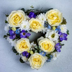 Sydämenmuotoinen kukkalaite, jossa on kermanvalkoisia ruusuja, liiloja freesioita ja sinisiä kukkia vihreällä pohjalla.