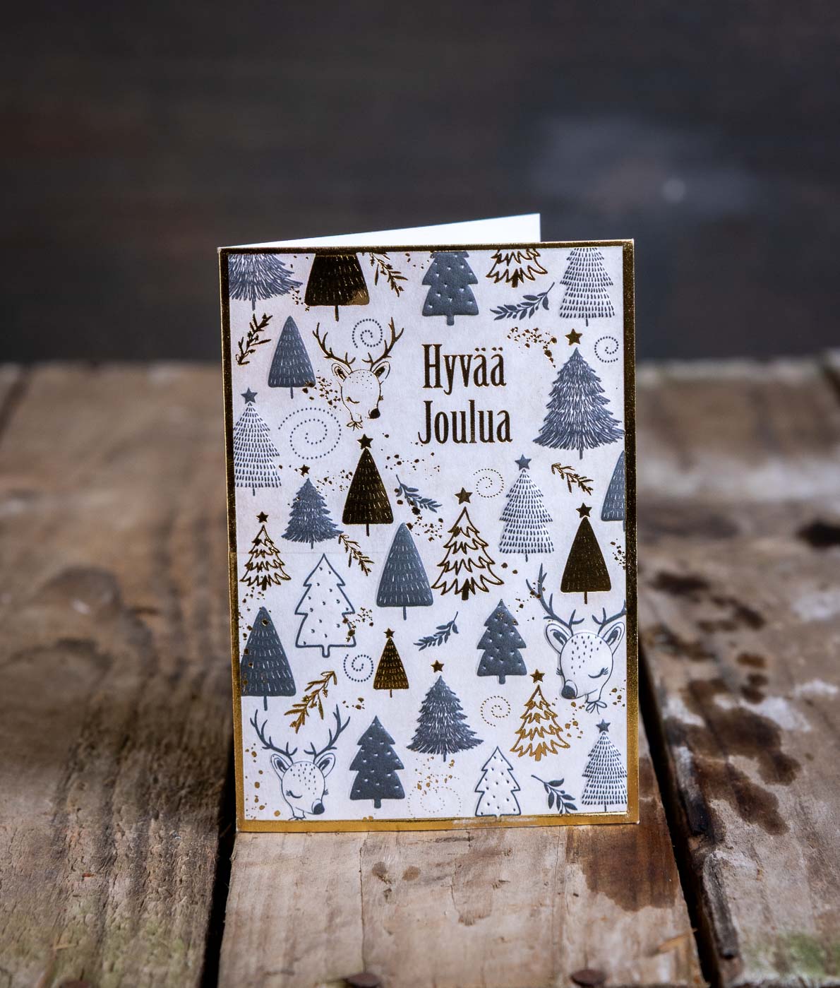 Pystyasennossa oleva joulukortti, jossa on kuvituksena erivärisiä joulukuusia, kultaisia yksityiskohtia, poron pää ja teksti Hyvää Joulua.