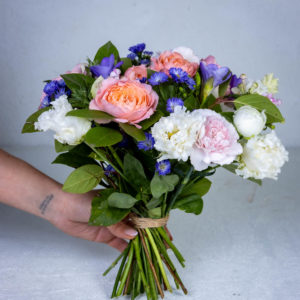 Runsas ja värikäs Kevään Kosketus -kukkakimppu, jossa on persikkaisia ruusuja ja violetteja kukkia.